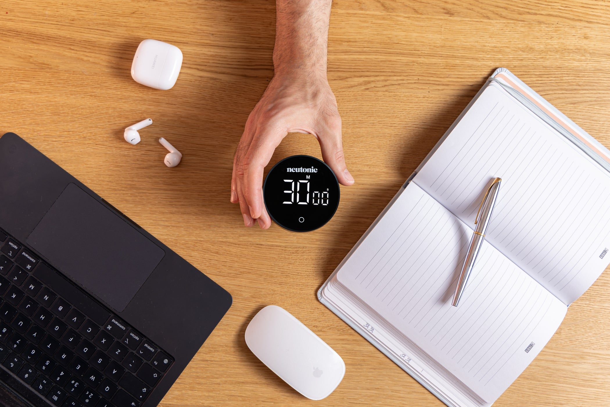Hand holding Focus Timer displaying 30 minutes on screen, surrounded by laptop, wireless earbuds, mouse, and open notebook. - Neutonic - Focus Timer